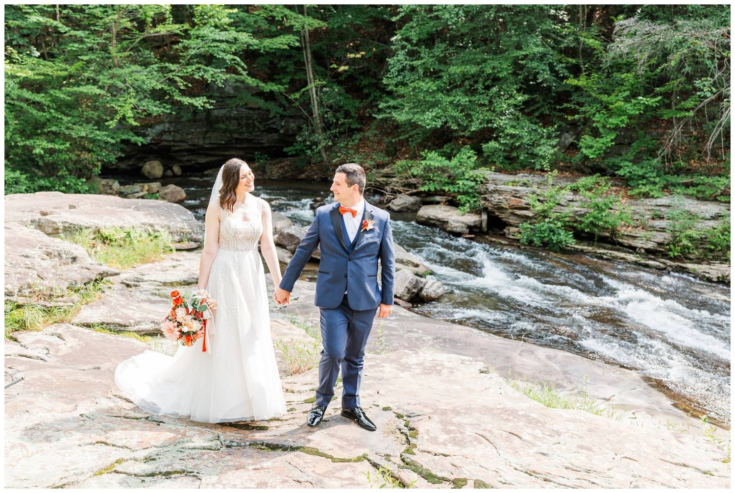 A couple stands together on rocky terrain near a flowing stream in a lush forest setting.