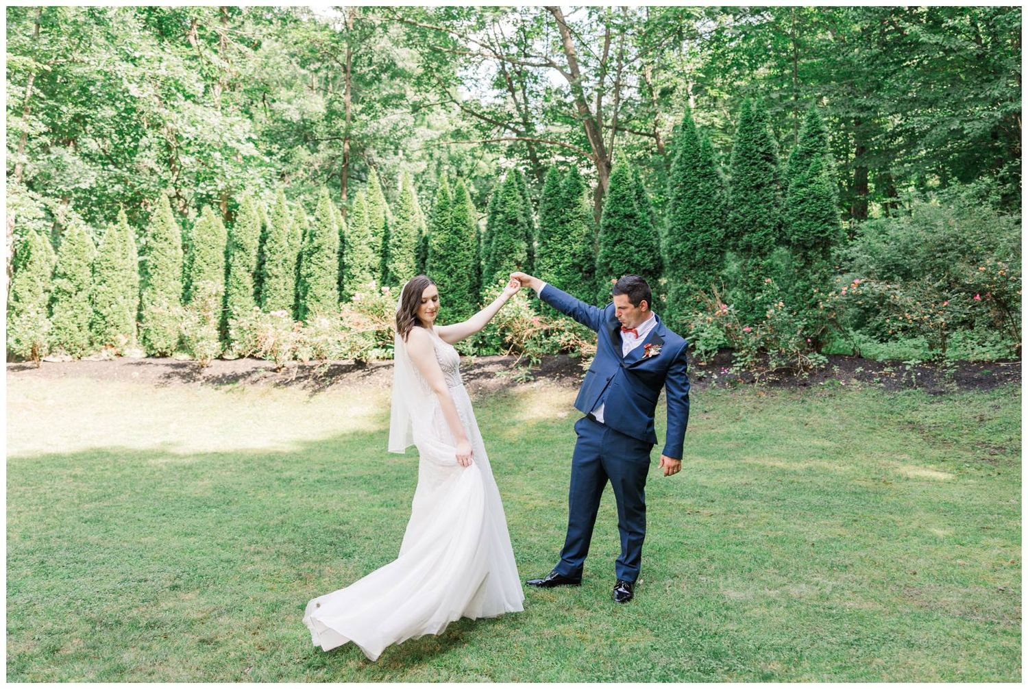 A couple dances together on a green lawn surrounded by evergreen trees on a bright summer day.