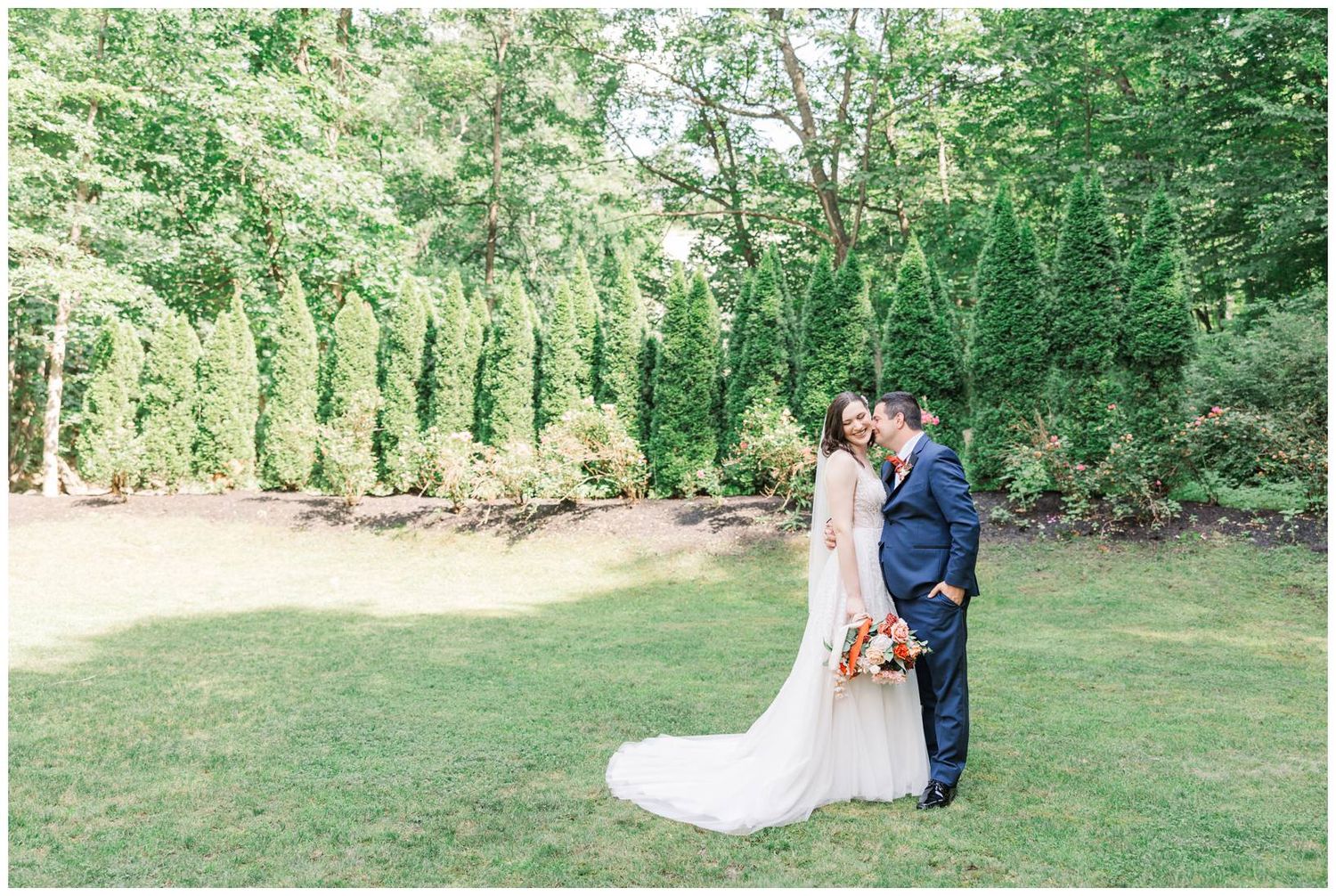 A bride in white gown and groom in navy suit embrace while standing on manicured lawn with evergreen backdrop.