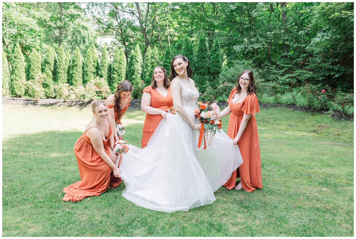 A bride in white gown poses outdoors with bridesmaids wearing coral-colored dresses on a green lawn surrounded by trees.