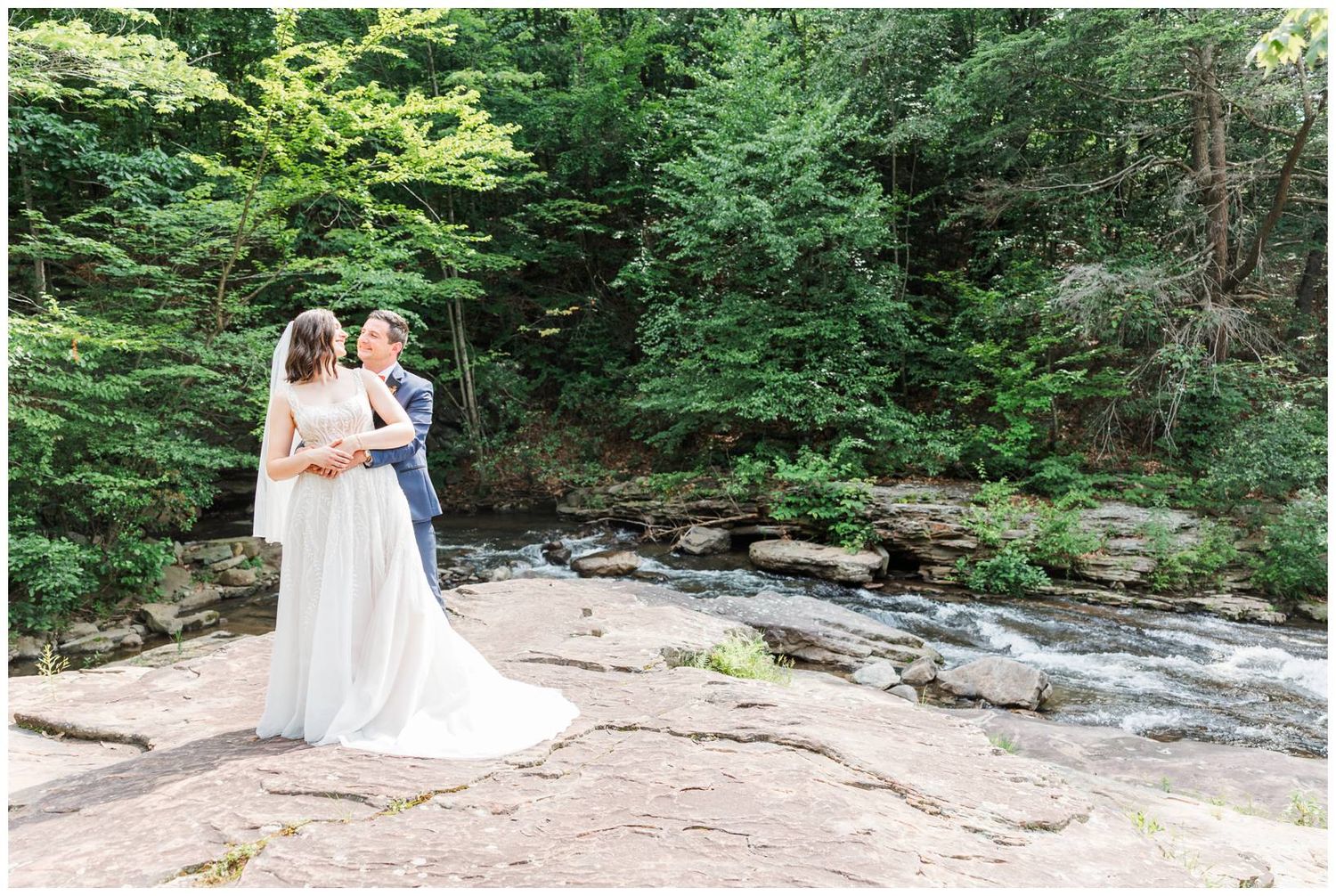 A couple in wedding attire stands on rocky terrain near a flowing stream surrounded by green forest.