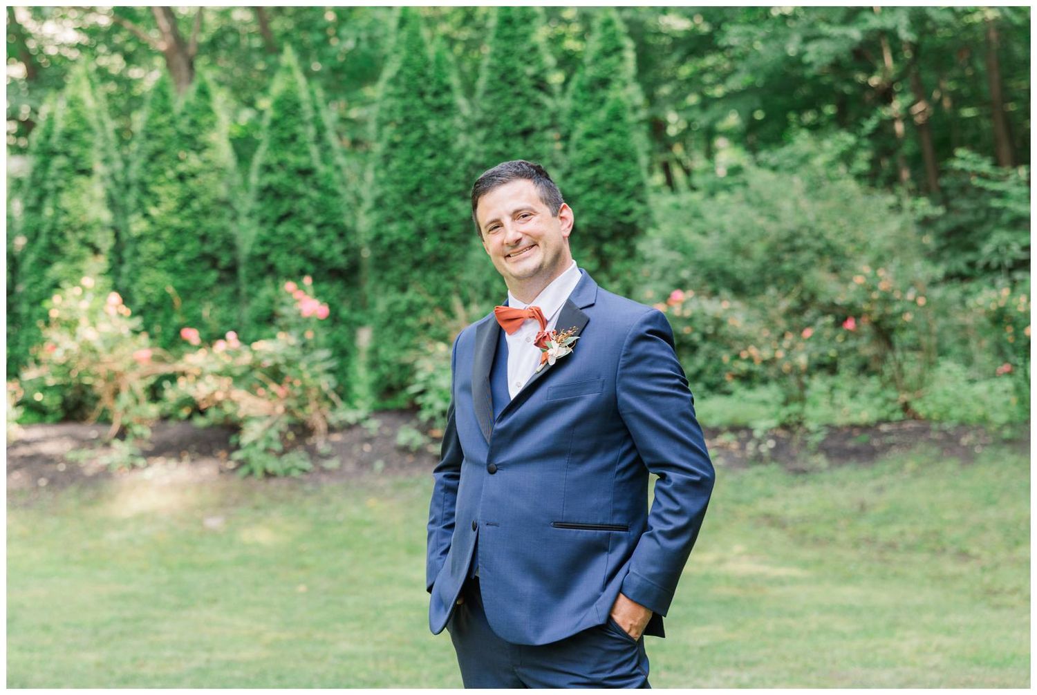 Wedding guest wearing a navy blue suit and coral boutonniere poses in a garden setting with evergreen trees in background.
