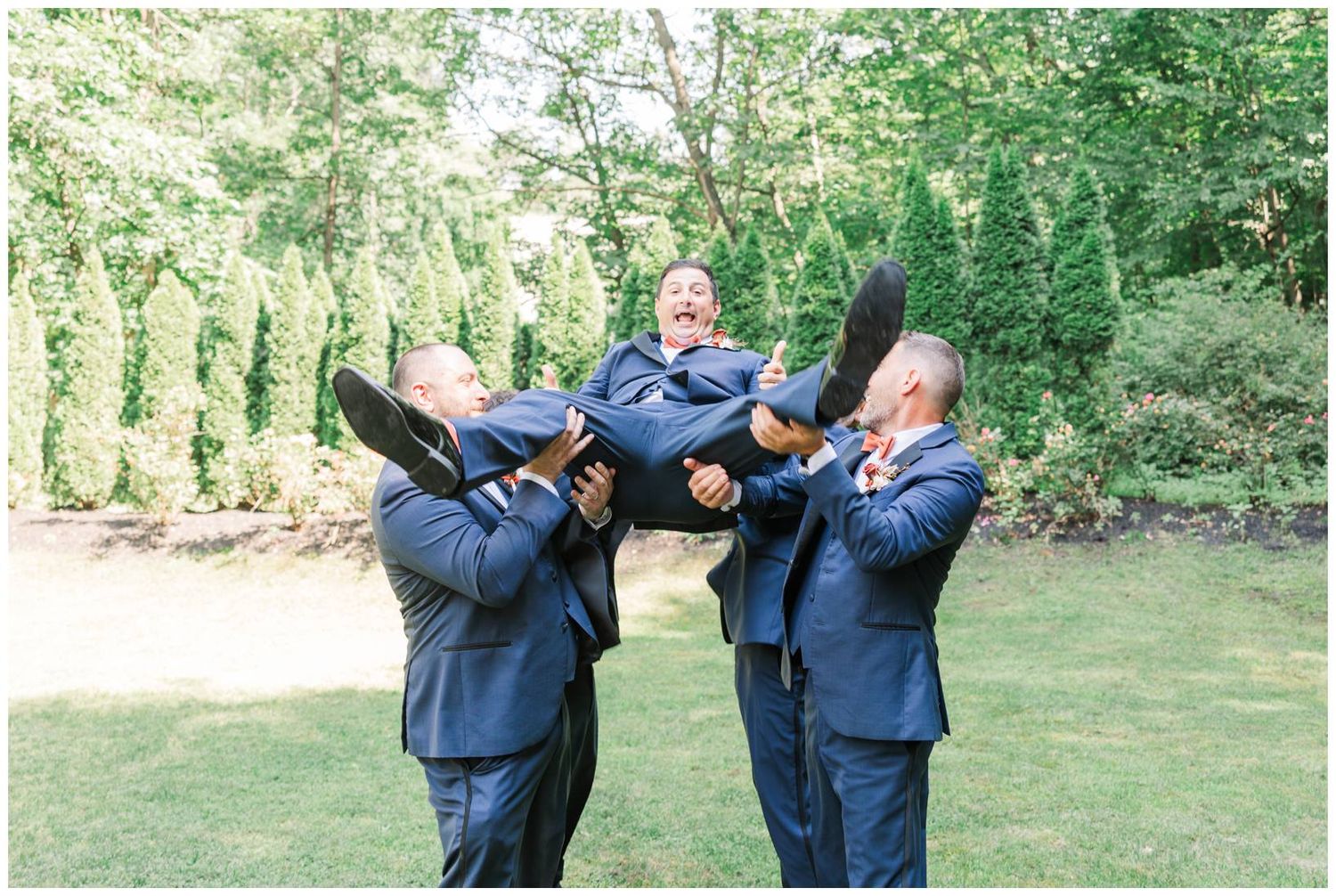 Three people in navy blue suits lift someone horizontally while standing on grass with green foliage in background.
