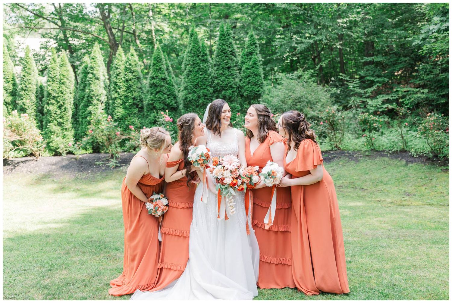 A bridal party wearing coral orange bridesmaid dresses poses outdoors in a lush green garden setting.
