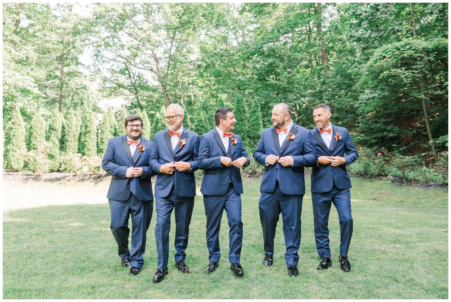 Groomsmen in navy suits walk playfully in a line across a grassy outdoor area at a wedding celebration.