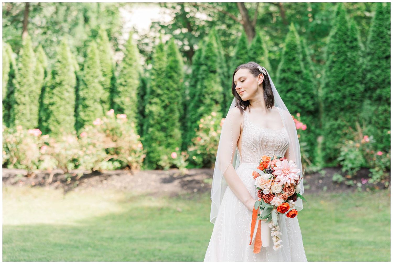 A bride in a flowing white gown poses outdoors holding a vibrant coral and peach floral bouquet in a garden setting.