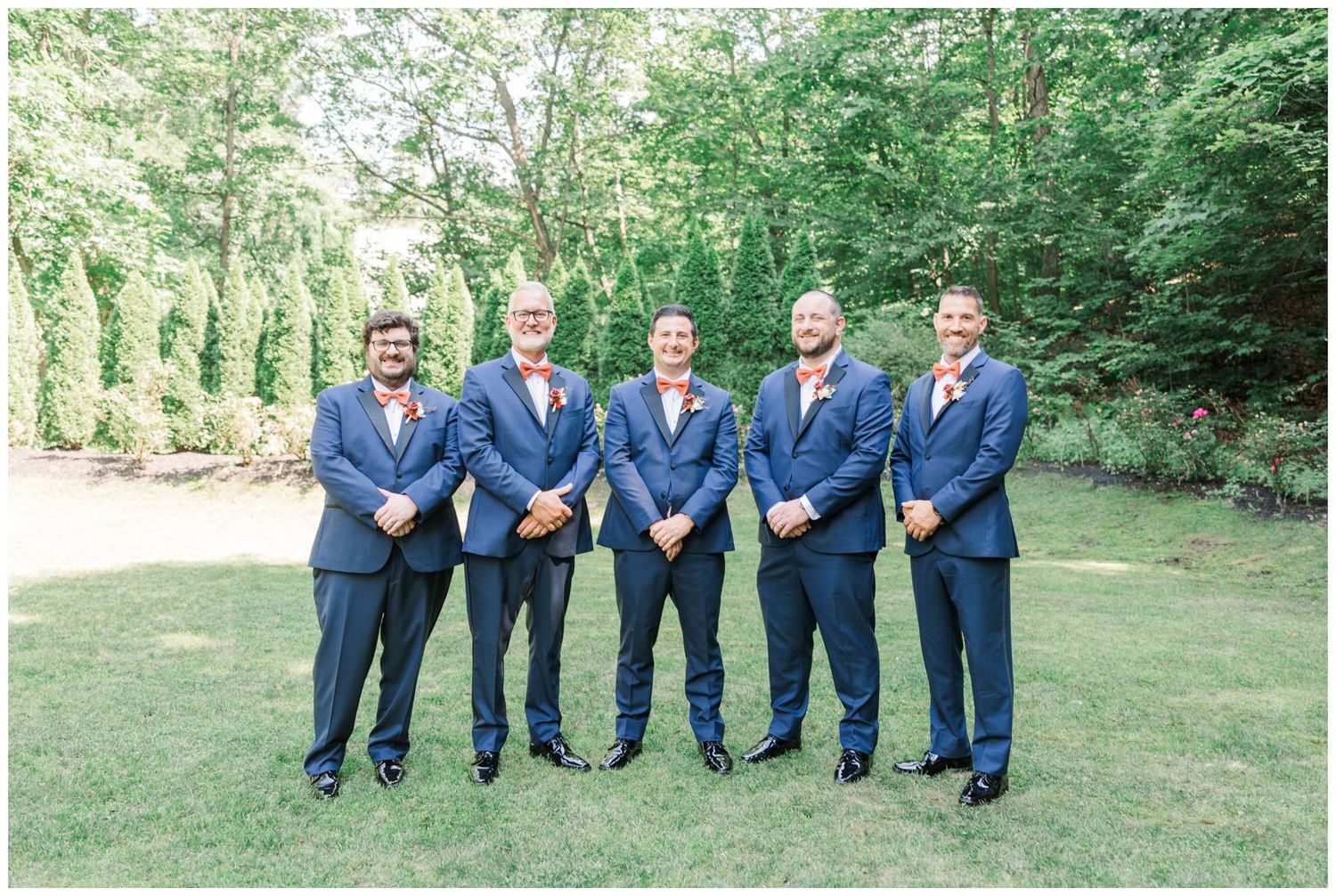Groomsmen in matching navy blue suits stand together on a grassy lawn surrounded by lush green trees.