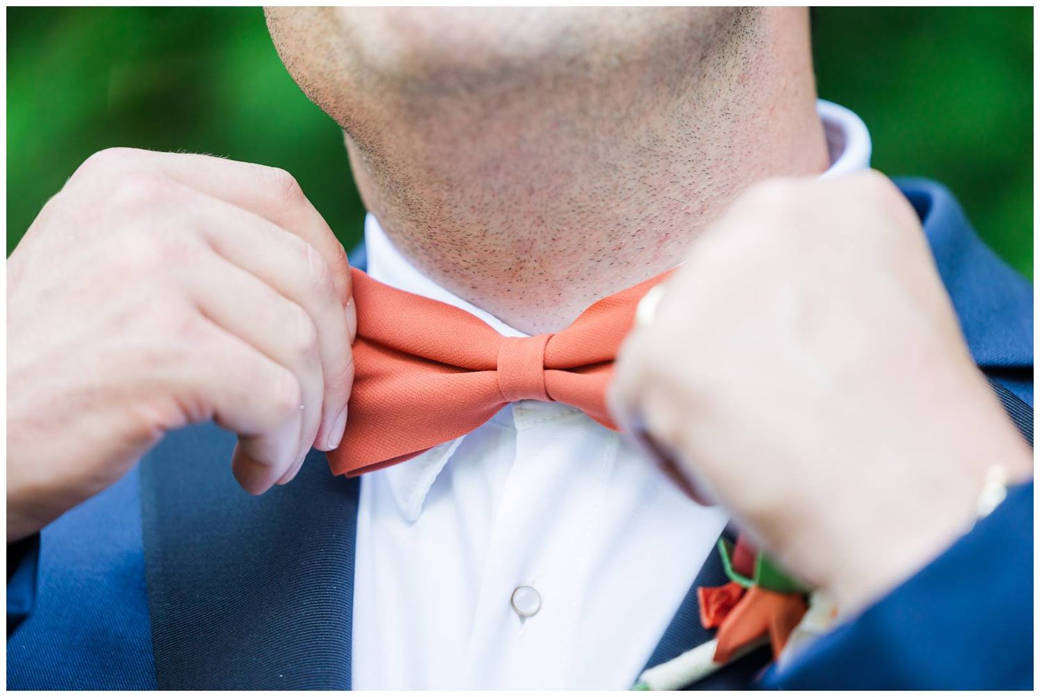 Hands adjusting a coral orange bow tie against a navy blue suit jacket.