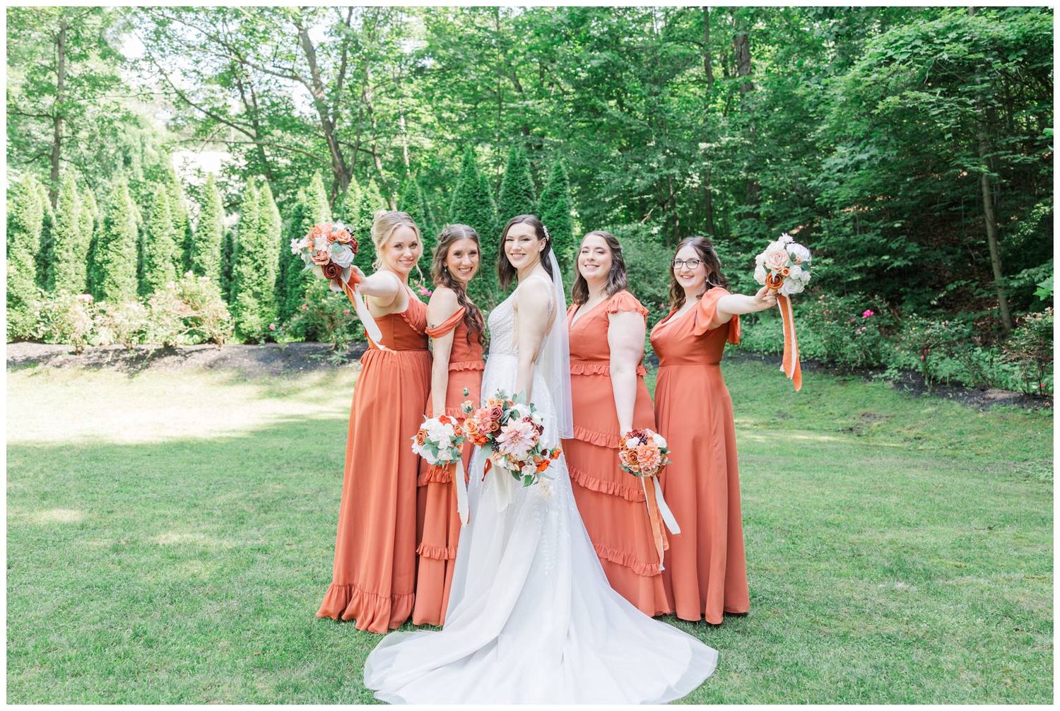 Bridal party in burnt orange dresses posing outdoors in natural garden setting.