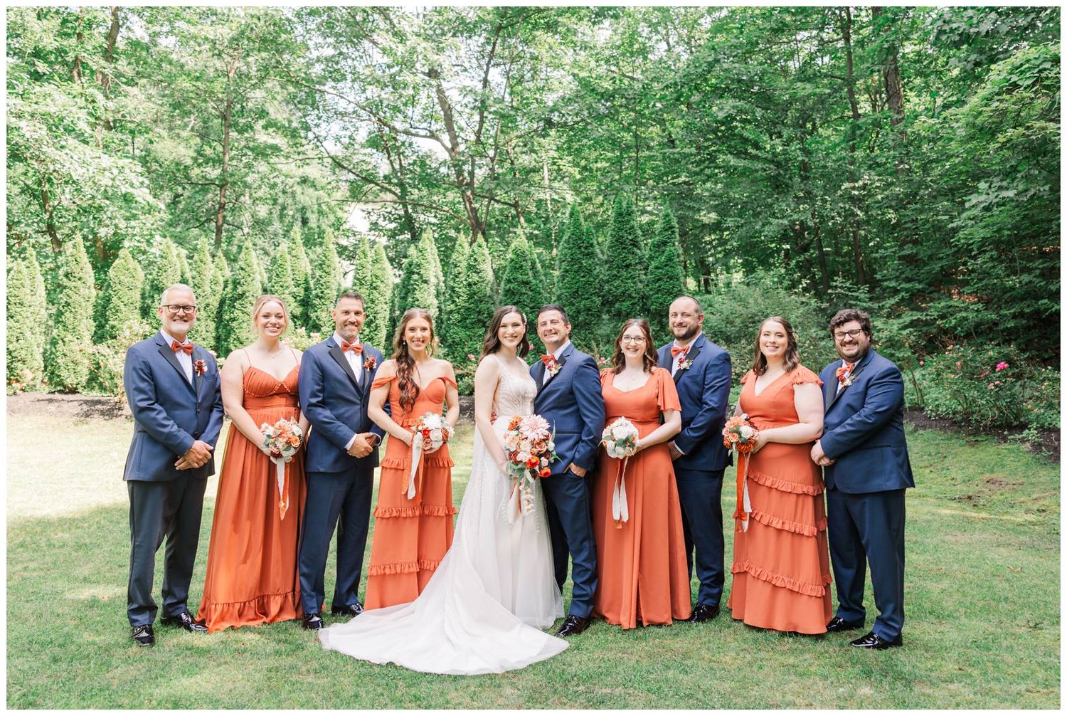 Wedding party in coral bridesmaids dresses and navy suits posed outdoors on lush green lawn.