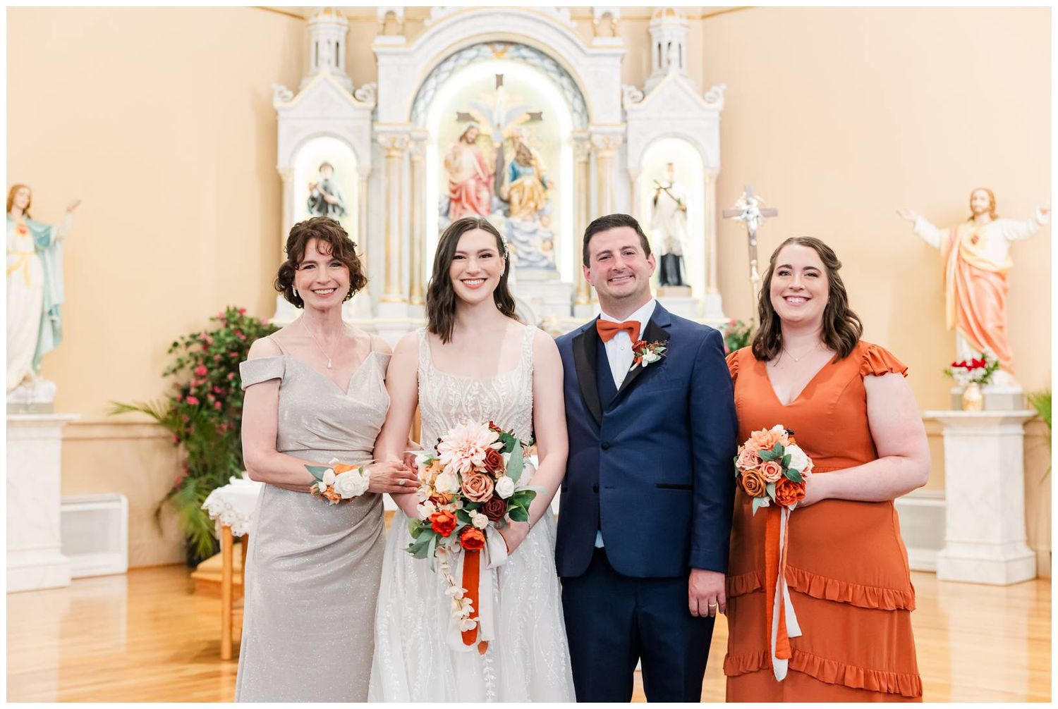 Wedding party in church wearing orange and navy attire standing near ornate altar.