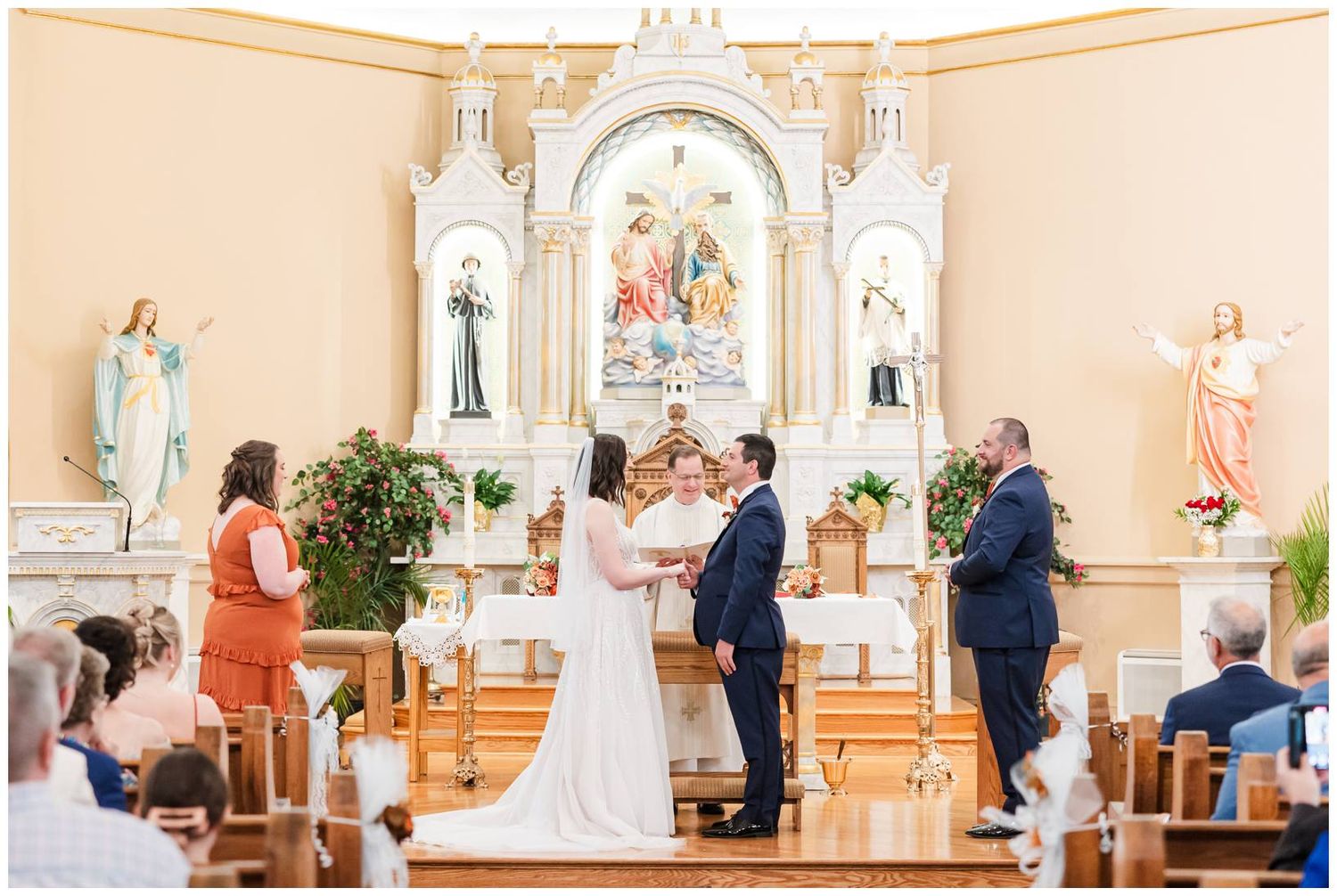 Traditional Catholic church wedding ceremony with elegant white decor and ornate altar in the background.