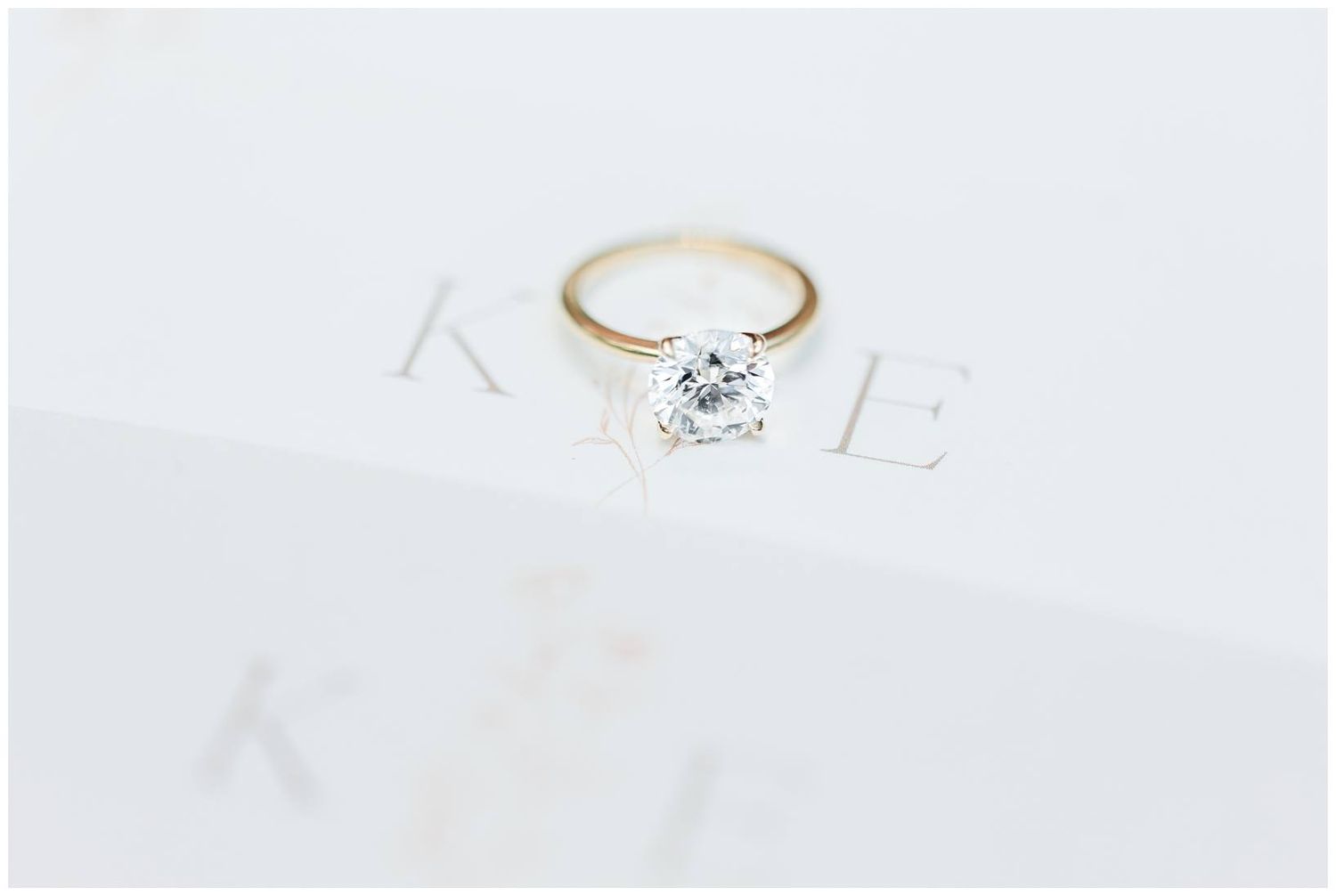 A delicate gold solitaire engagement ring with a round diamond sits atop a white textured surface.