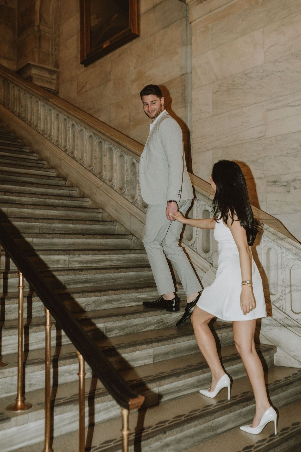 Couple poses on grand marble staircase in elegant formal attire during engagement photoshoot.