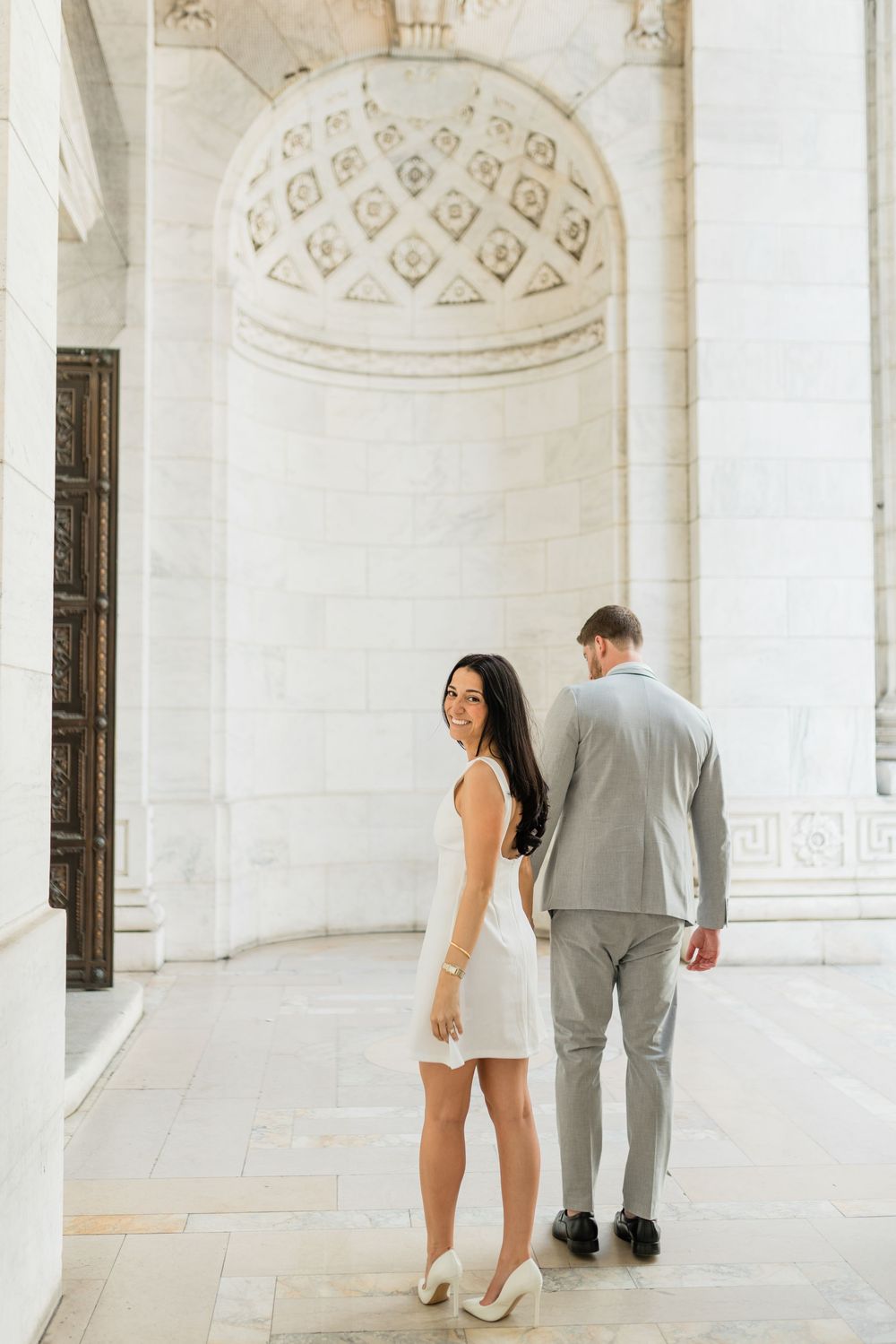A couple in light colored formal wear poses in an elegant architectural setting with decorative dome.