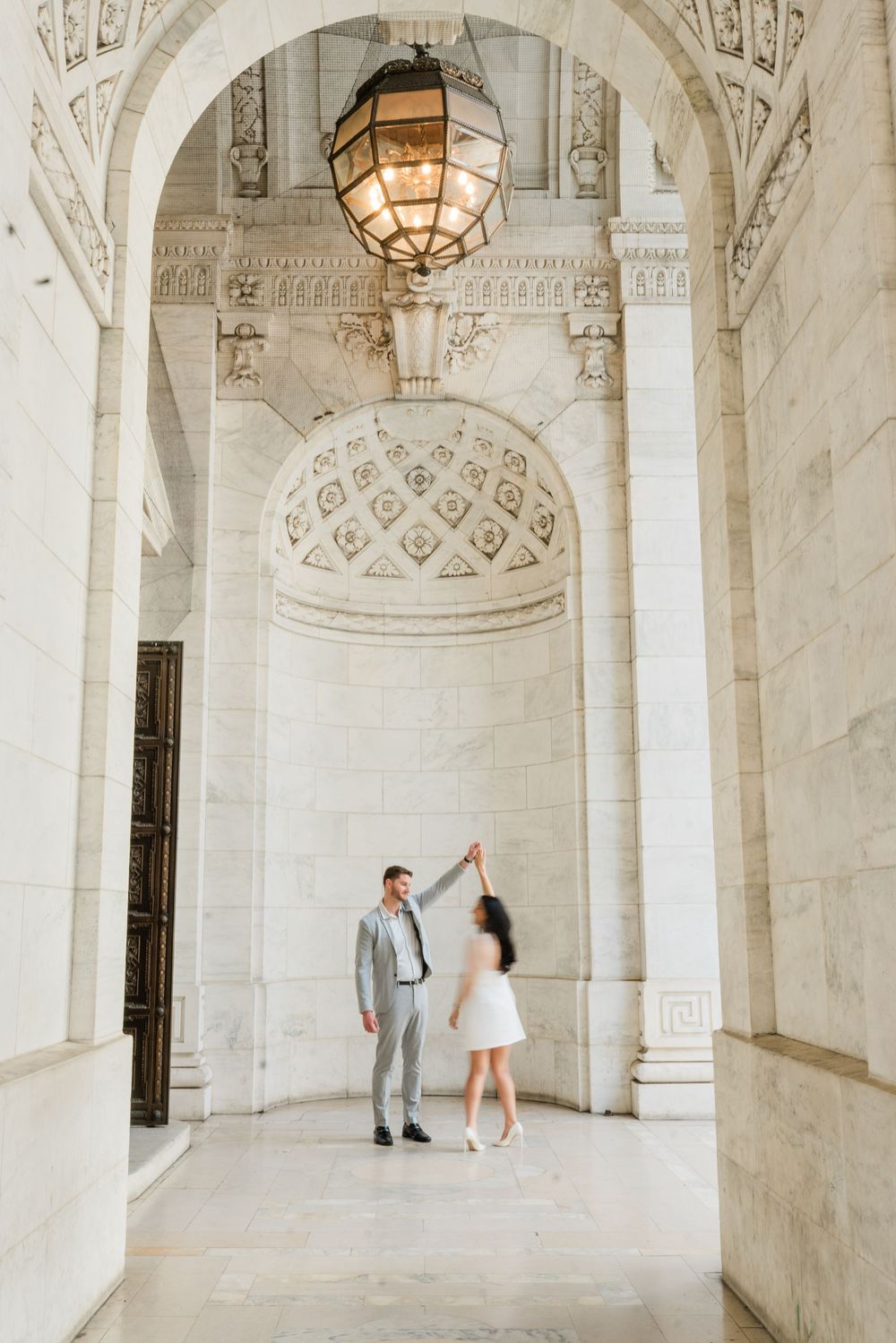 A couple dances beneath a grand hanging lantern in a classical architectural hallway.