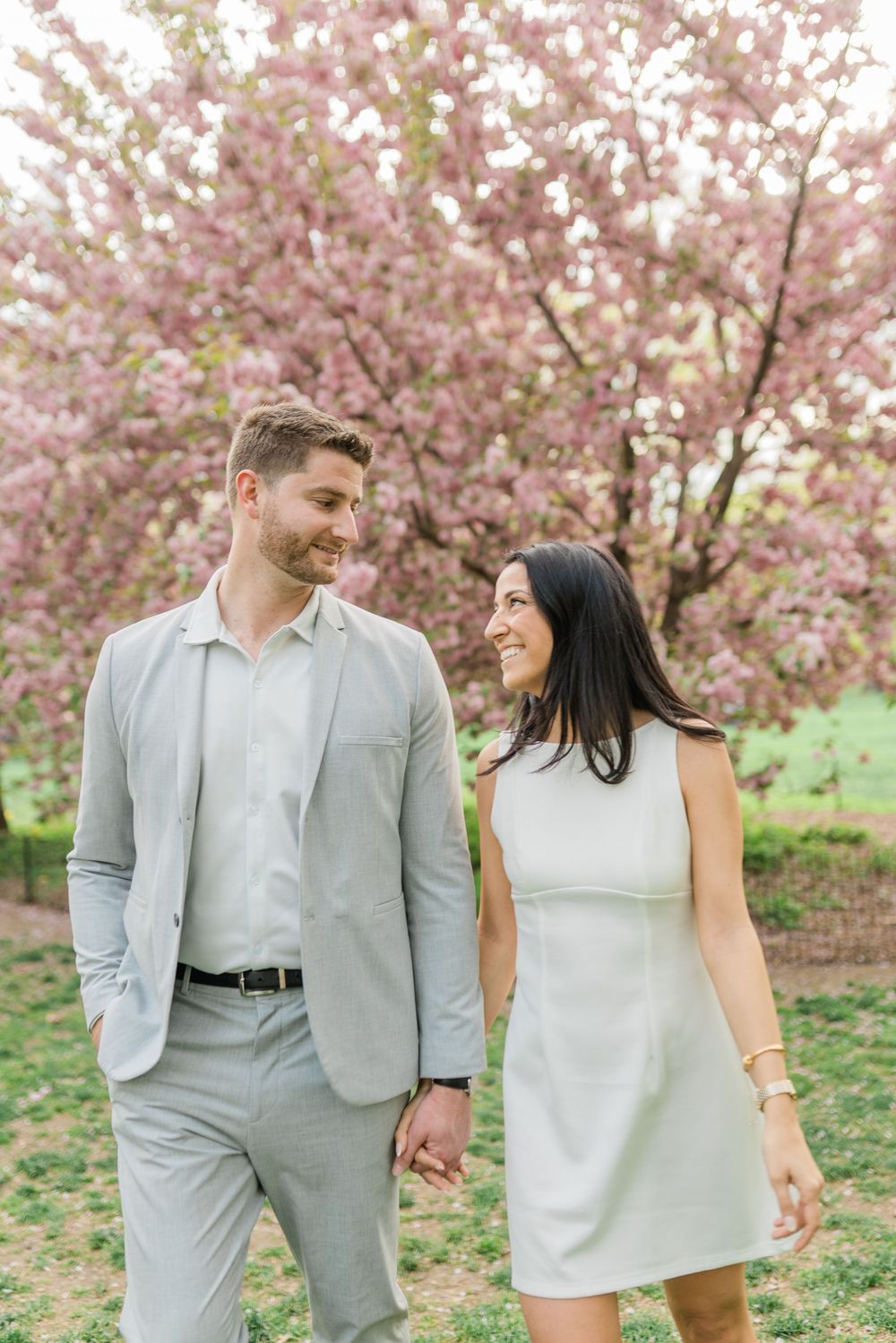 A couple in coordinating light outfits walks beneath blooming pink cherry blossoms.