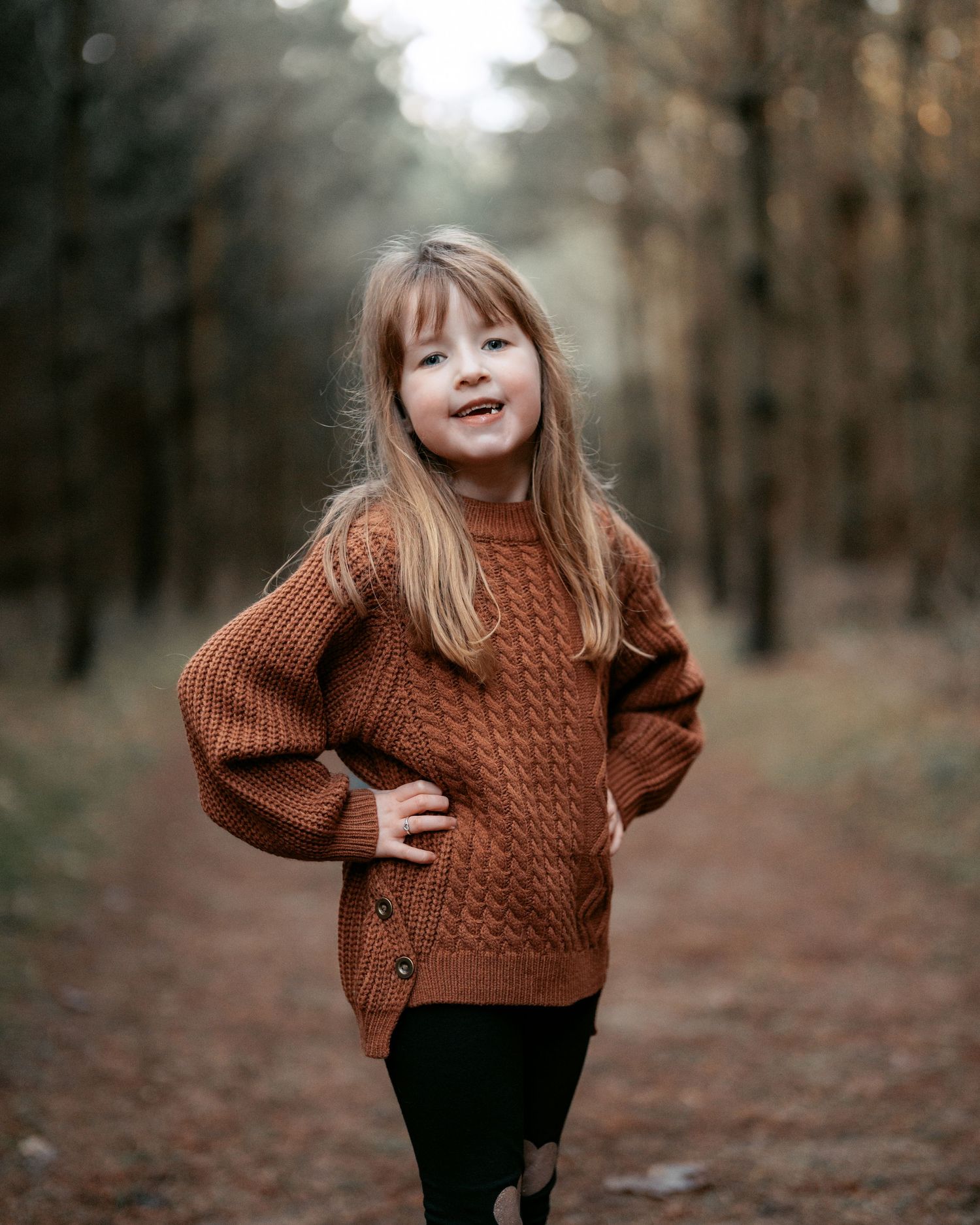 Young child wearing brown cable knit sweater poses playfully on autumn forest path surrounded by trees.