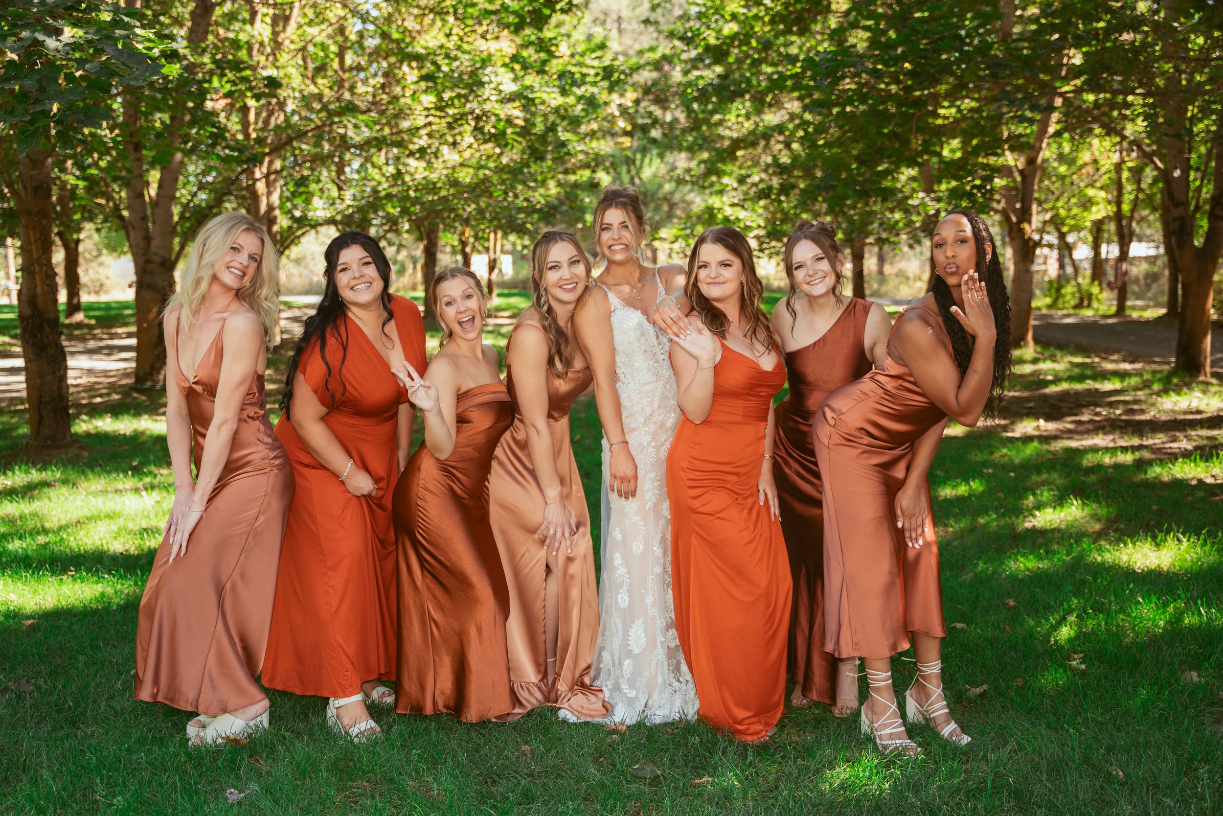 Group of bridesmaids in coordinating rust and terracotta colored dresses pose together on green grass in a park.