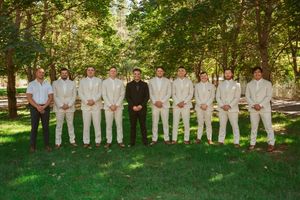 Wedding party wearing white suits poses with groomsmen in a sunny garden setting.