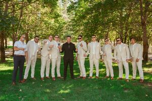 Groomsmen in light colored suits stand with person in black on grass under trees.