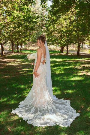 A bride in a stunning lace wedding gown with a flowing train poses in a sunlit garden setting.