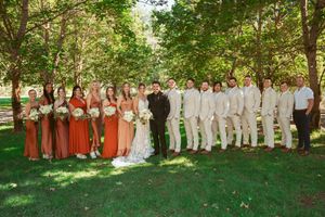 Wedding party with bridesmaids in coral dresses and groomsmen in white suits pose together on lawn.
