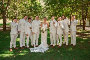 Wedding party wearing all-white attire poses playfully together for group photos in a sunny garden setting.