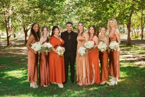 Wedding party members in rust-colored dresses and dark attire standing together outdoors.