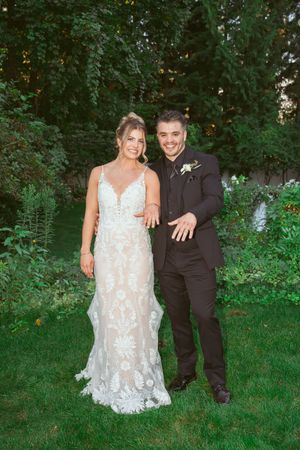 A couple poses in a garden setting with the bride wearing a lace white gown and groom in black suit.