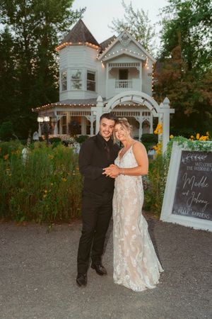 A newlywed couple stands in front of a charming Victorian house decorated with string lights.