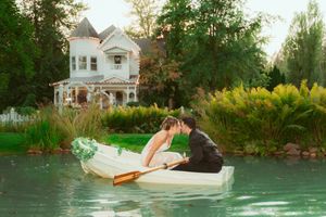 A romantic couple in wedding attire sits in a rowboat on a pond with a Victorian mansion in background.