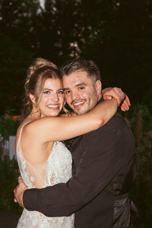 A couple embraces and smiles while wearing wedding attire during an outdoor celebration.