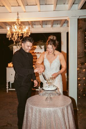 Couple cutting their wedding cake together under elegant chandelier in rustic venue.
