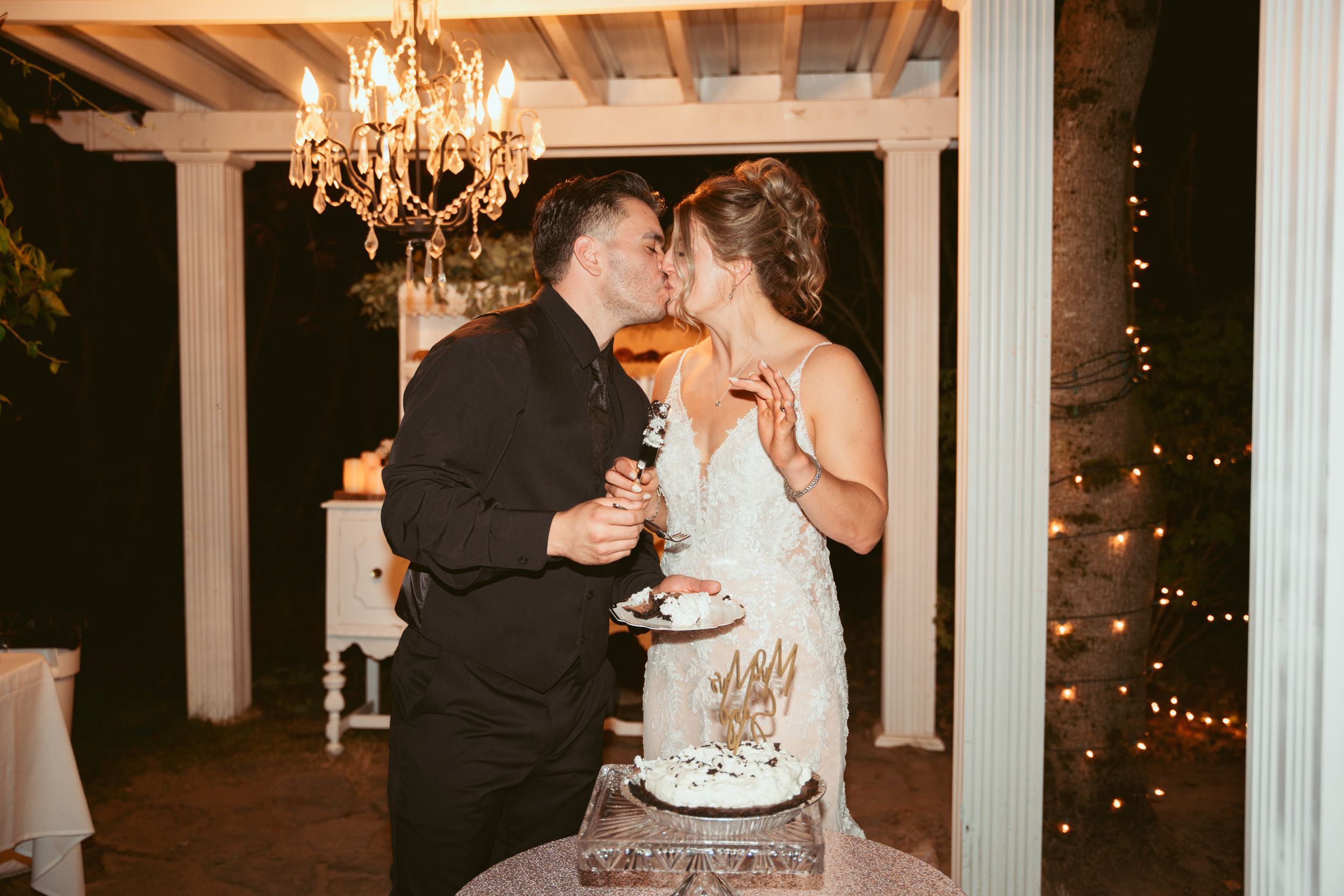 Newlyweds share cake and champagne under chandelier at outdoor reception.