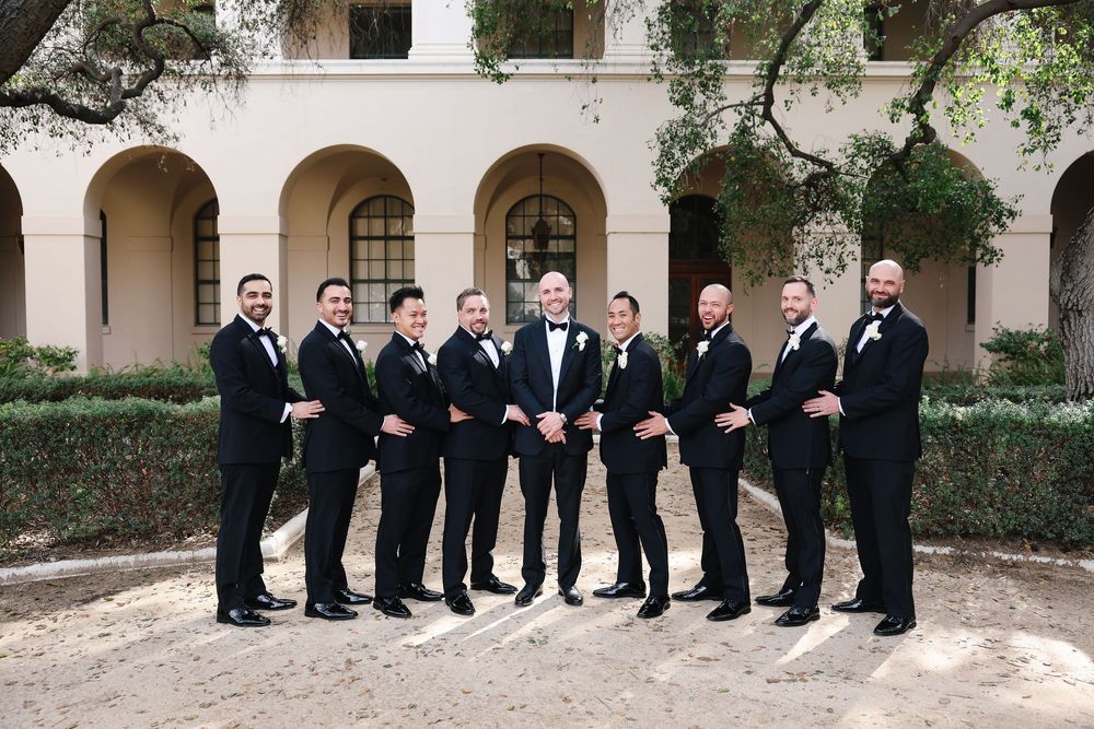 A wedding party in matching black tuxedos stands in a row at an elegant venue with archways and pink walls.