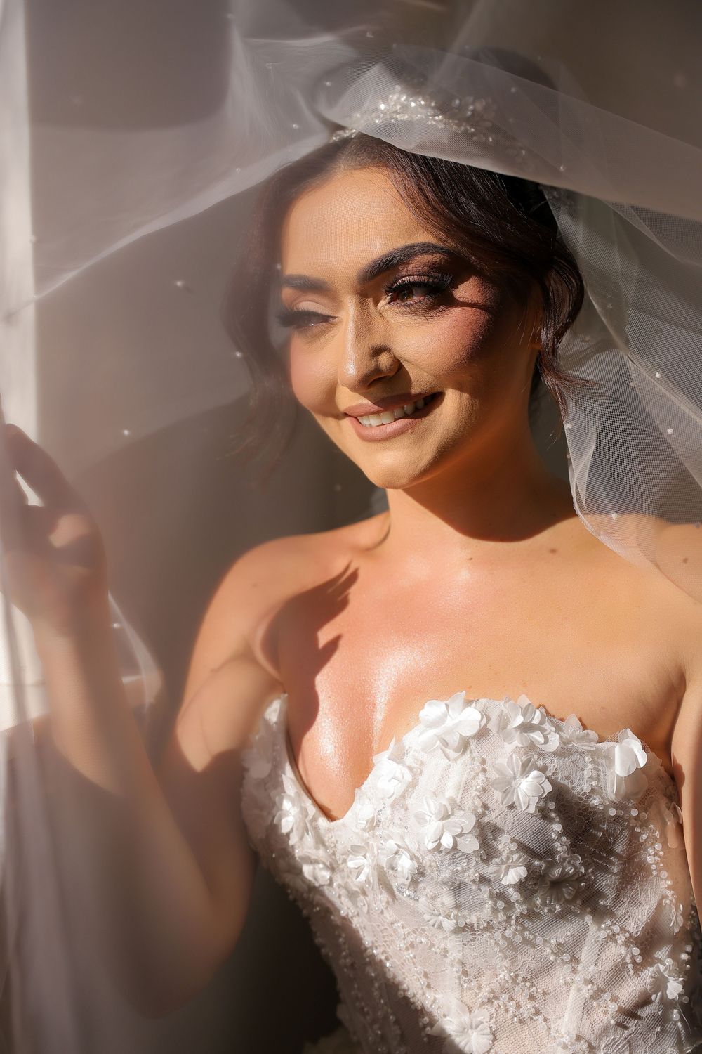A bride wearing a strapless white wedding gown with delicate lace details and a flowing veil poses in soft lighting.