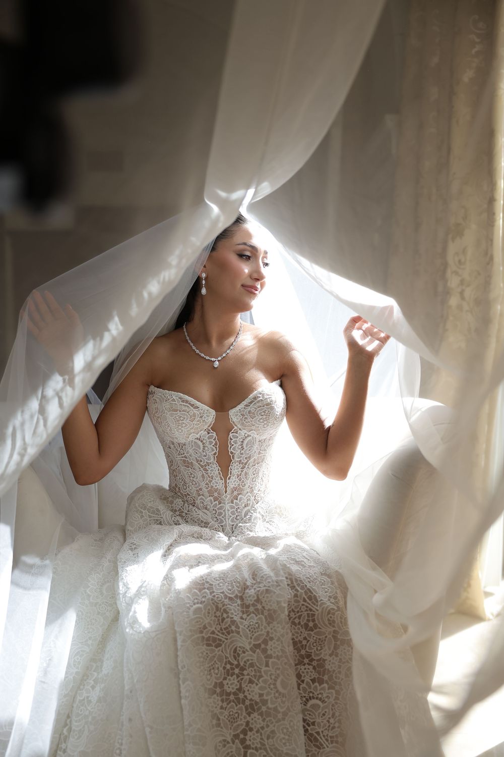 A bride in an elegant lace wedding dress poses with her flowing veil in soft natural window light.