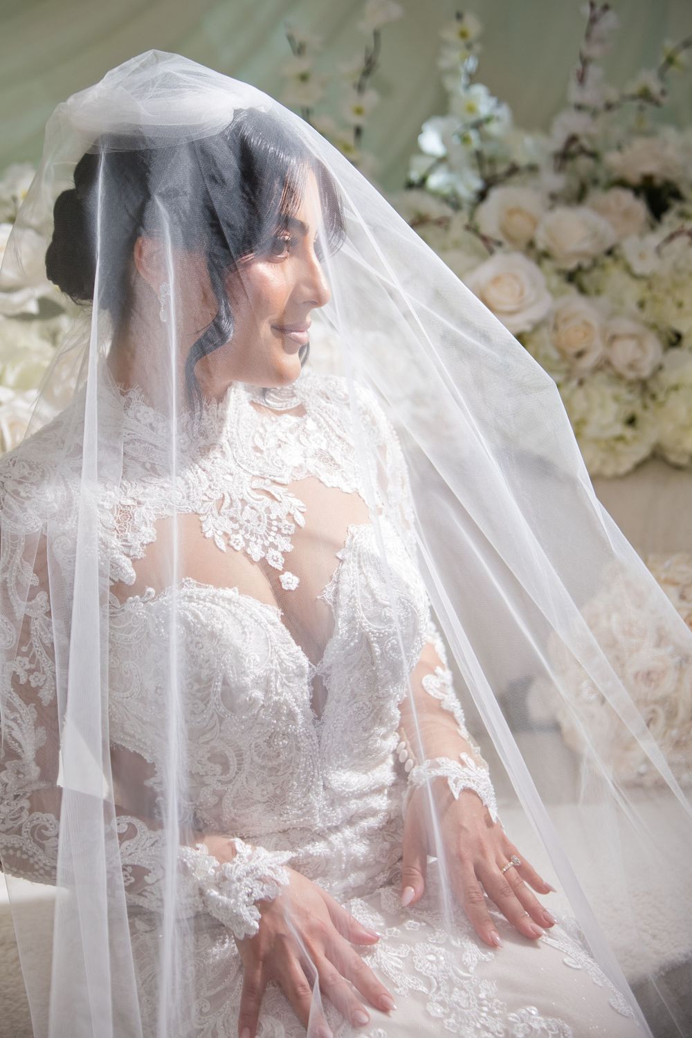 A bride in a delicate lace wedding dress sits gracefully behind a flowing white veil creating an ethereal bridal portrait.