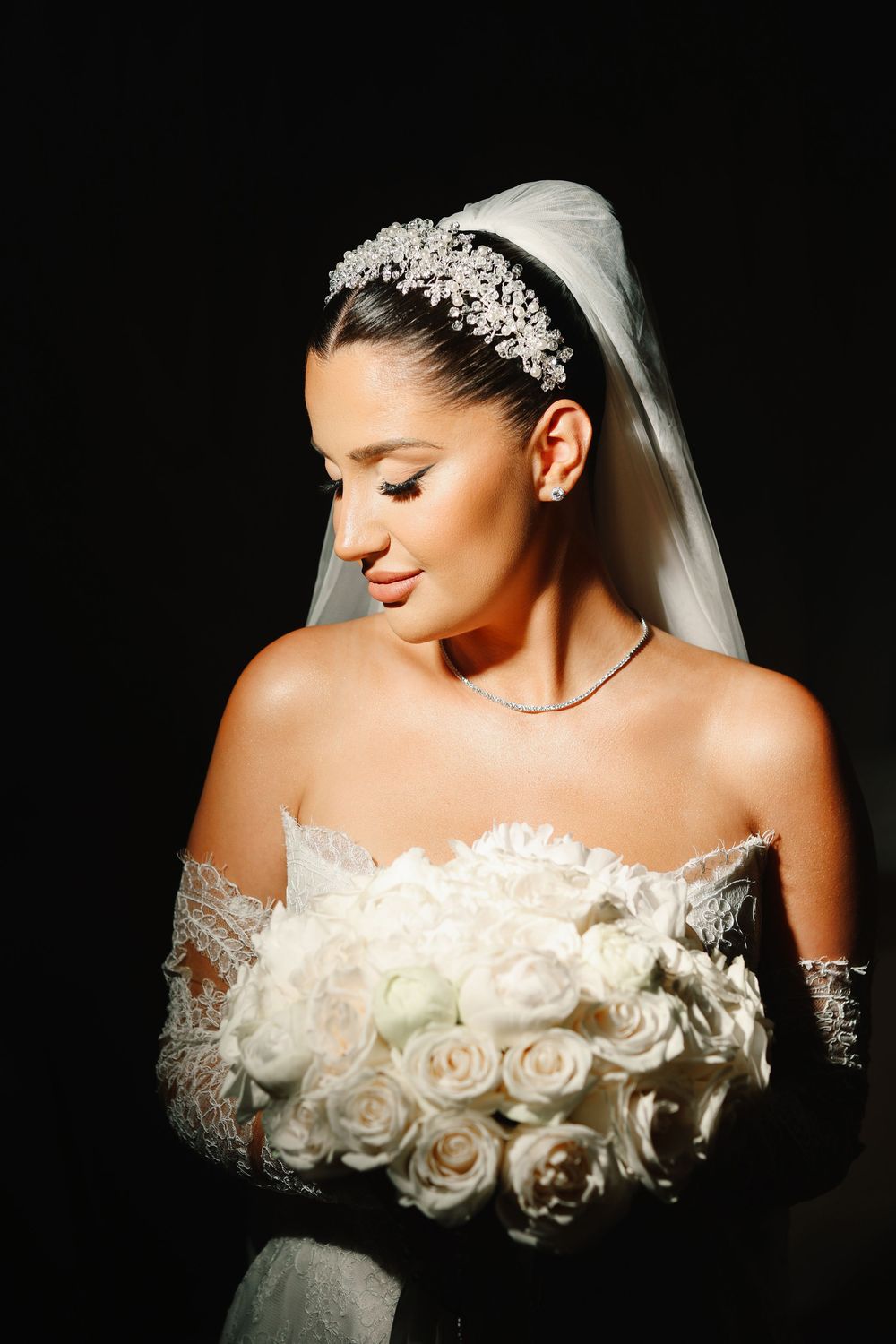 Wedding portrait showing a close-up view of a bride wearing an off-shoulder rose-adorned gown and delicate veil against dark backdrop.