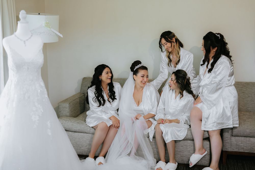 Bridal party in white robes with her bride made relaxing on gray couch with wedding dress displayed nearby.