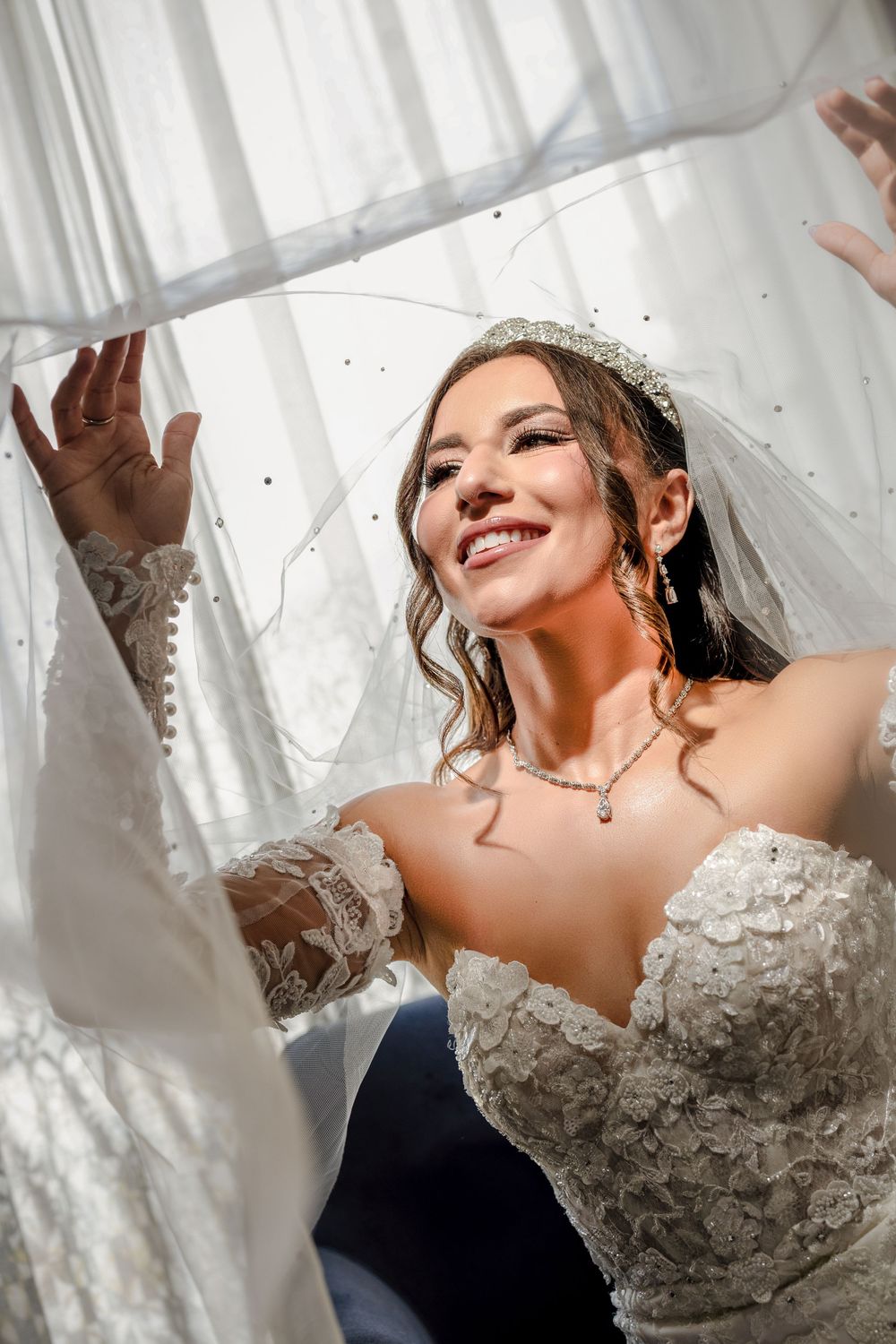 Beautiful bride in lace wedding dress and veil smiling joyfully in natural window light.