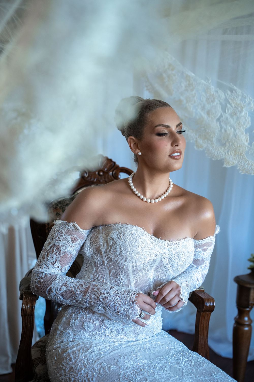 Elegant off-shoulder lace wedding dress with pearl necklace and flowing veil in dreamy ethereal setting.