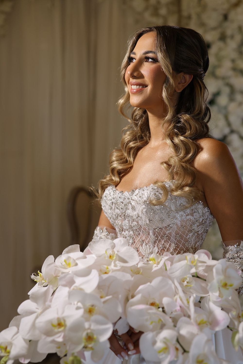Elegant bride in lace dress poses next to stunning white orchid bouquet in natural window light.