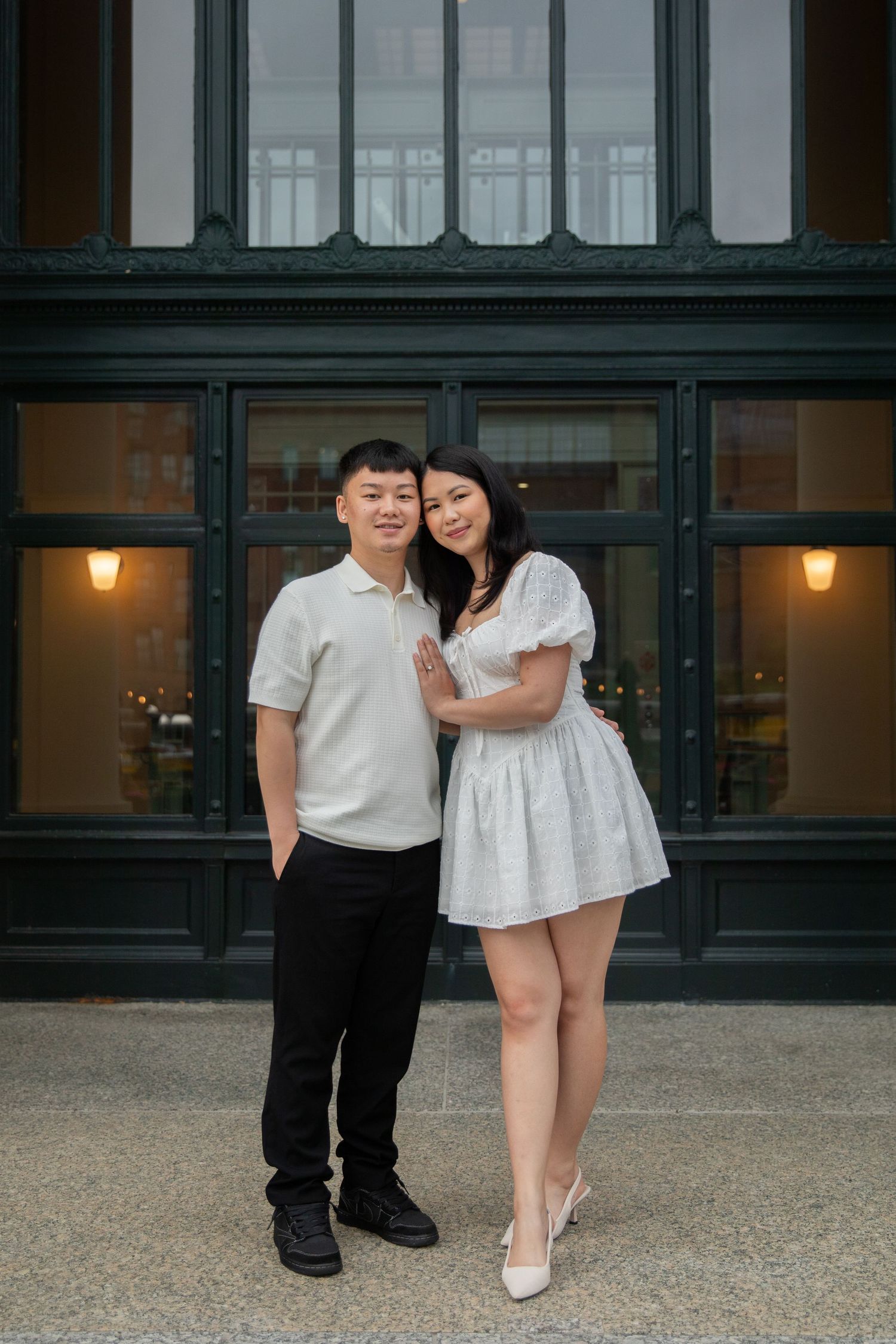A couple in coordinated white outfits poses together in front of a dark storefront with warm lighting.
