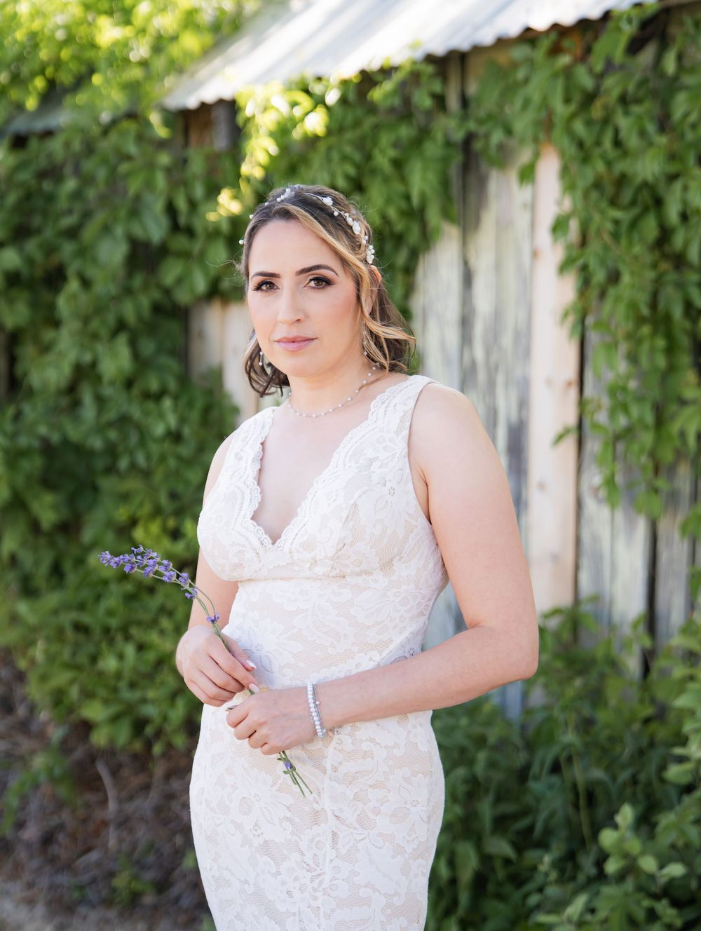 Beautiful bride in white lace dress holding lavender sprig poses outdoors against rustic barn wall with climbing vines.