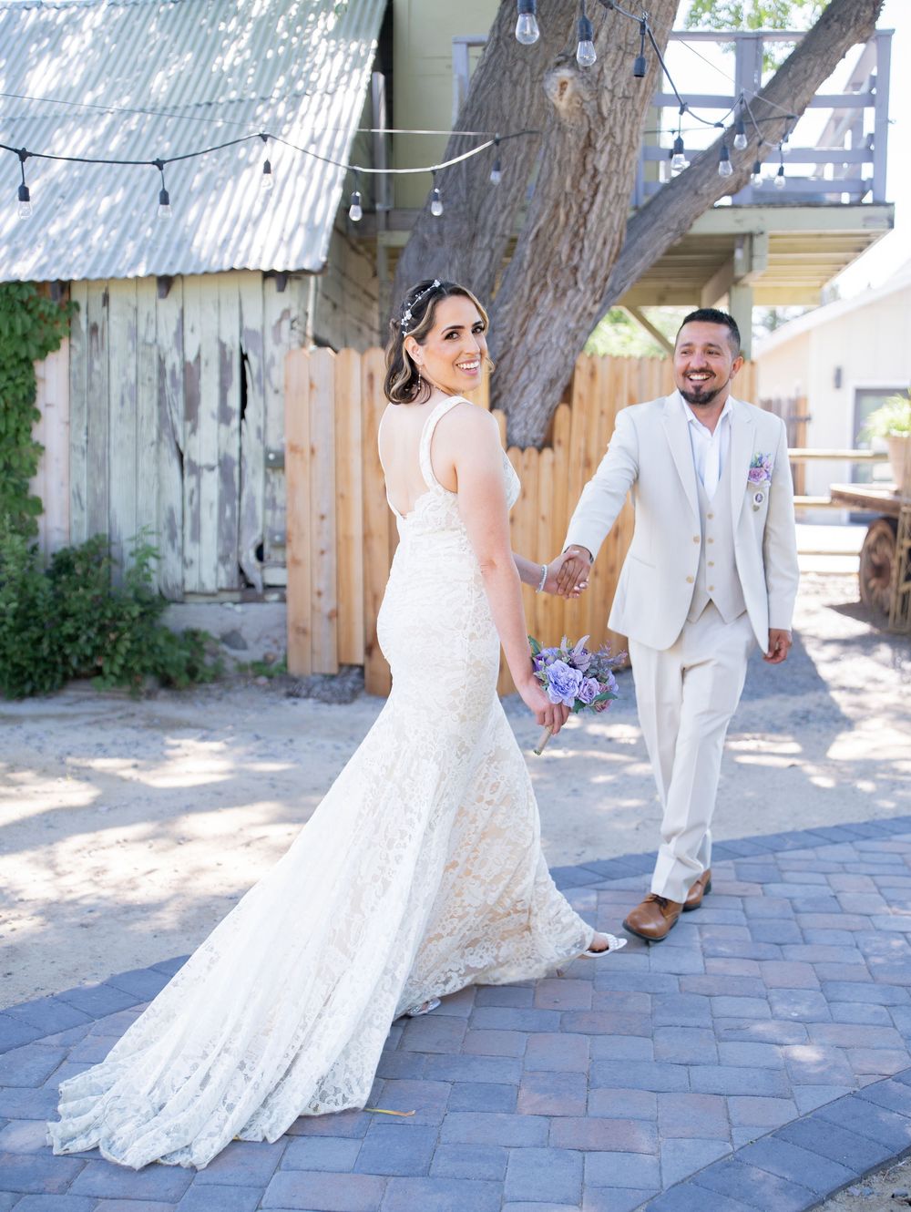 A joyful couple on their wedding day walking on brick pavement with rustic barn and string lights in background.