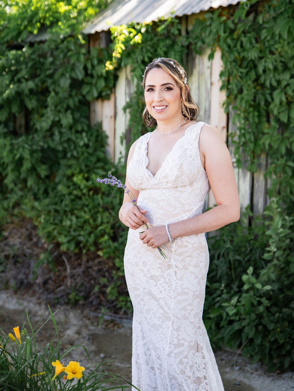 A bride in a white lace wedding gown poses against an ivy-covered wall while holding a lavender sprig on a sunny day.