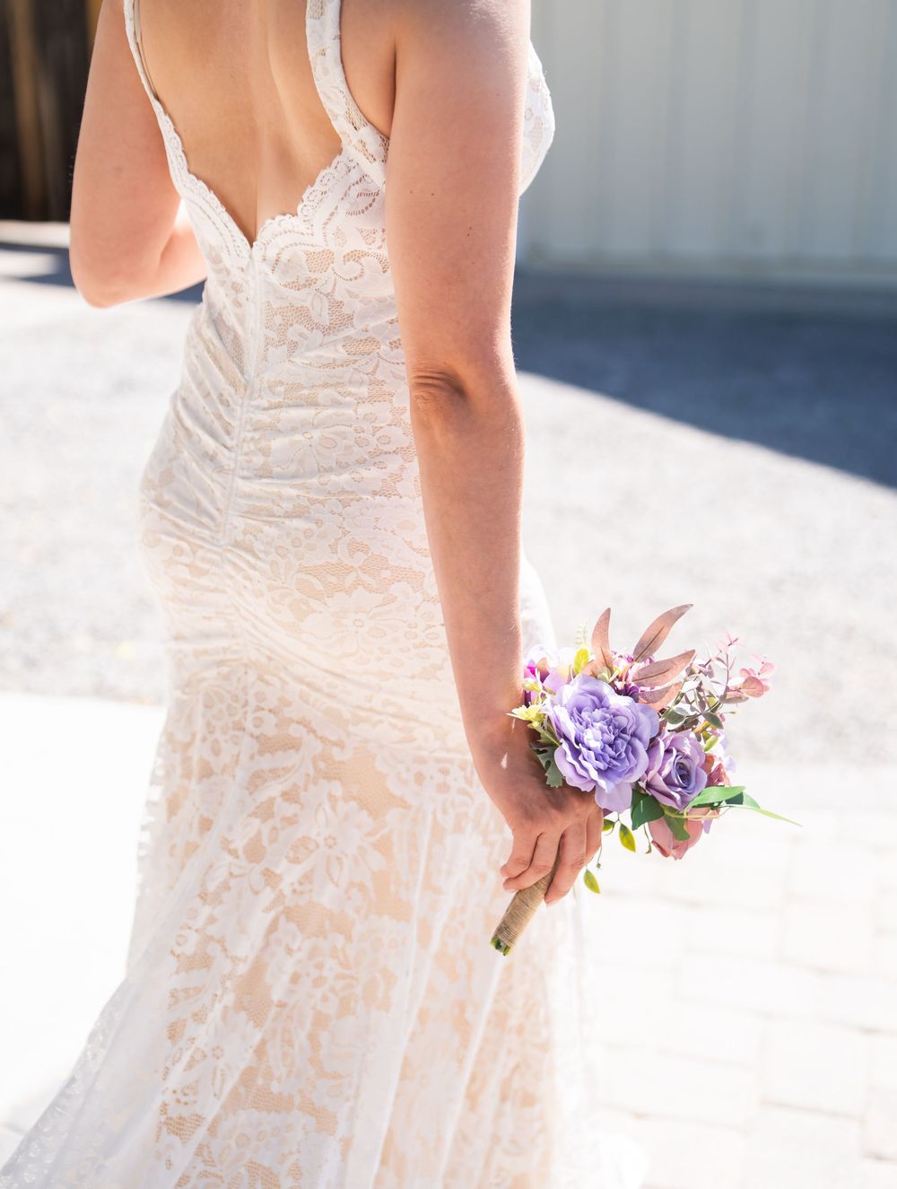 Elegant lace wedding dress details shown in close-up with delicate floral bouquet of purple and pink blooms in sunlight.