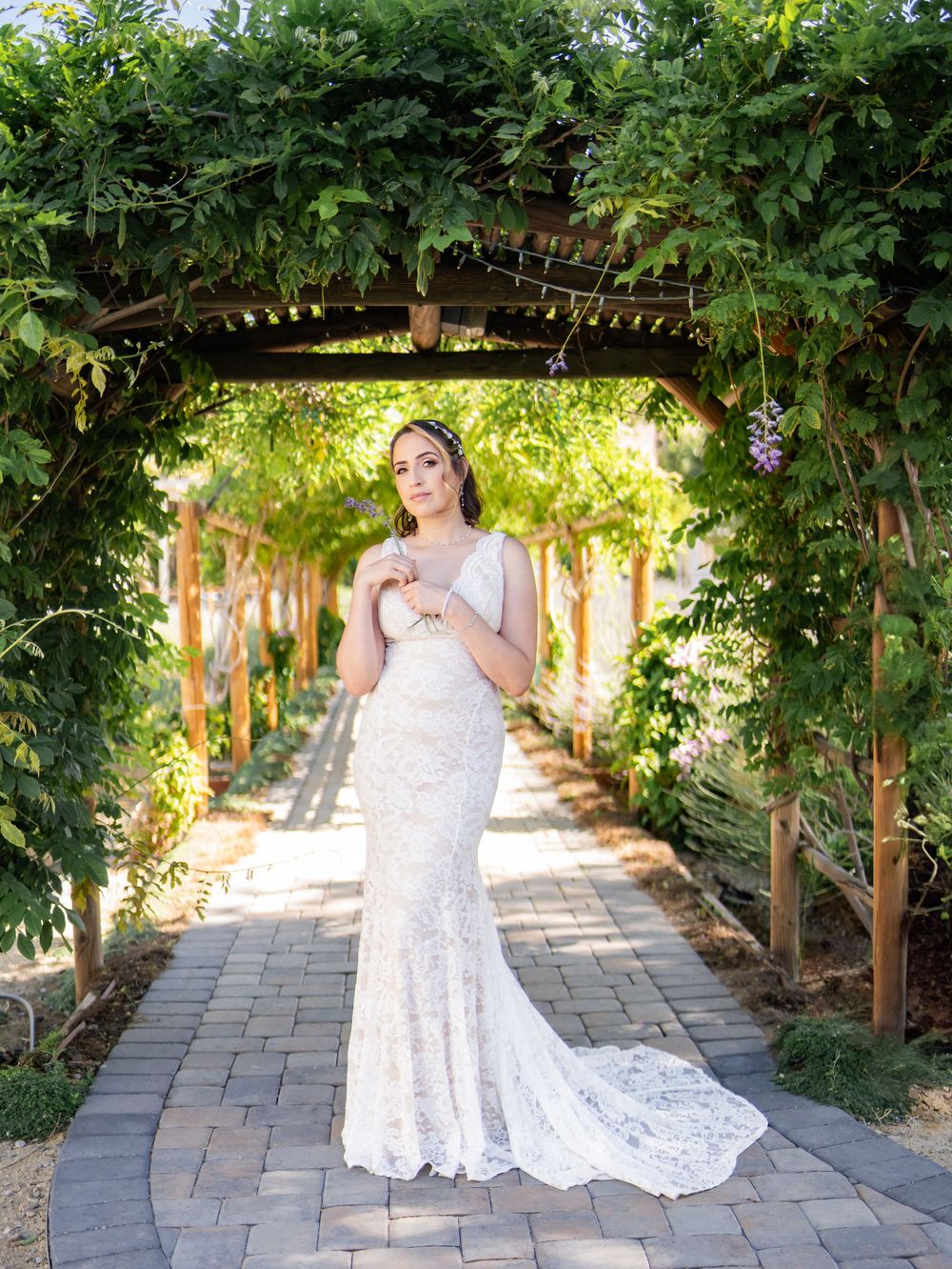 A bride in a flowing lace wedding gown stands beneath a vine-covered garden archway on a stone pathway.